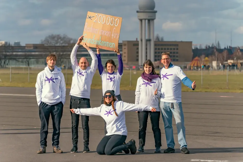 Die Mitglieder der Expedition Grundeinkommen stehen auf dem Tempelhofer Feld und halten ein Plakat mit der Aufschrift "200.000 Berliner innen fürs bedingungslose Grundeinkommen